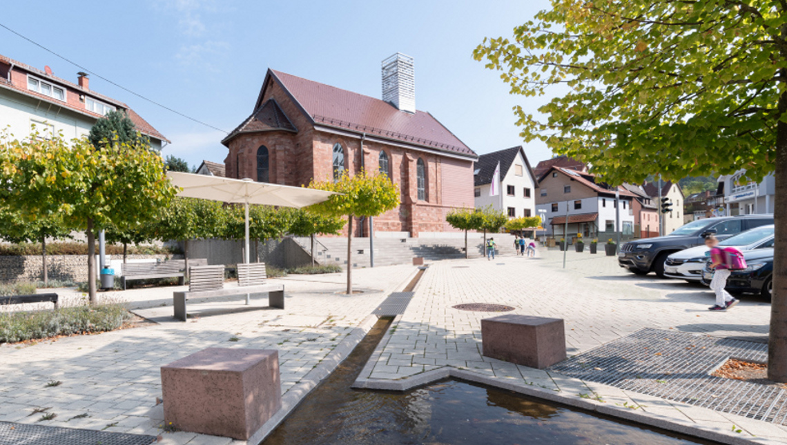 Hell gepflasterter Dorfplatz in Altenbach mit jungen Bäumen und Kirche im Hintergrund